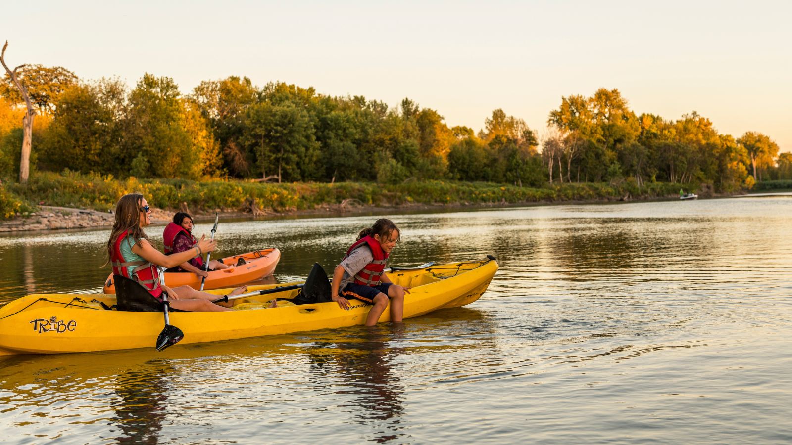 A group of kayakers on the Red River in Grand Forks kayak along during golden hour. One kid hangs off the side to dip their feet in the water.