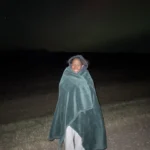 Author Precious stands and smiles at the camera during a northern lights chasing trip while she's wrapped up in a cozy blanket!