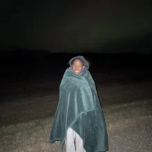 Author Precious stands and smiles at the camera during a northern lights chasing trip while she's wrapped up in a cozy blanket!